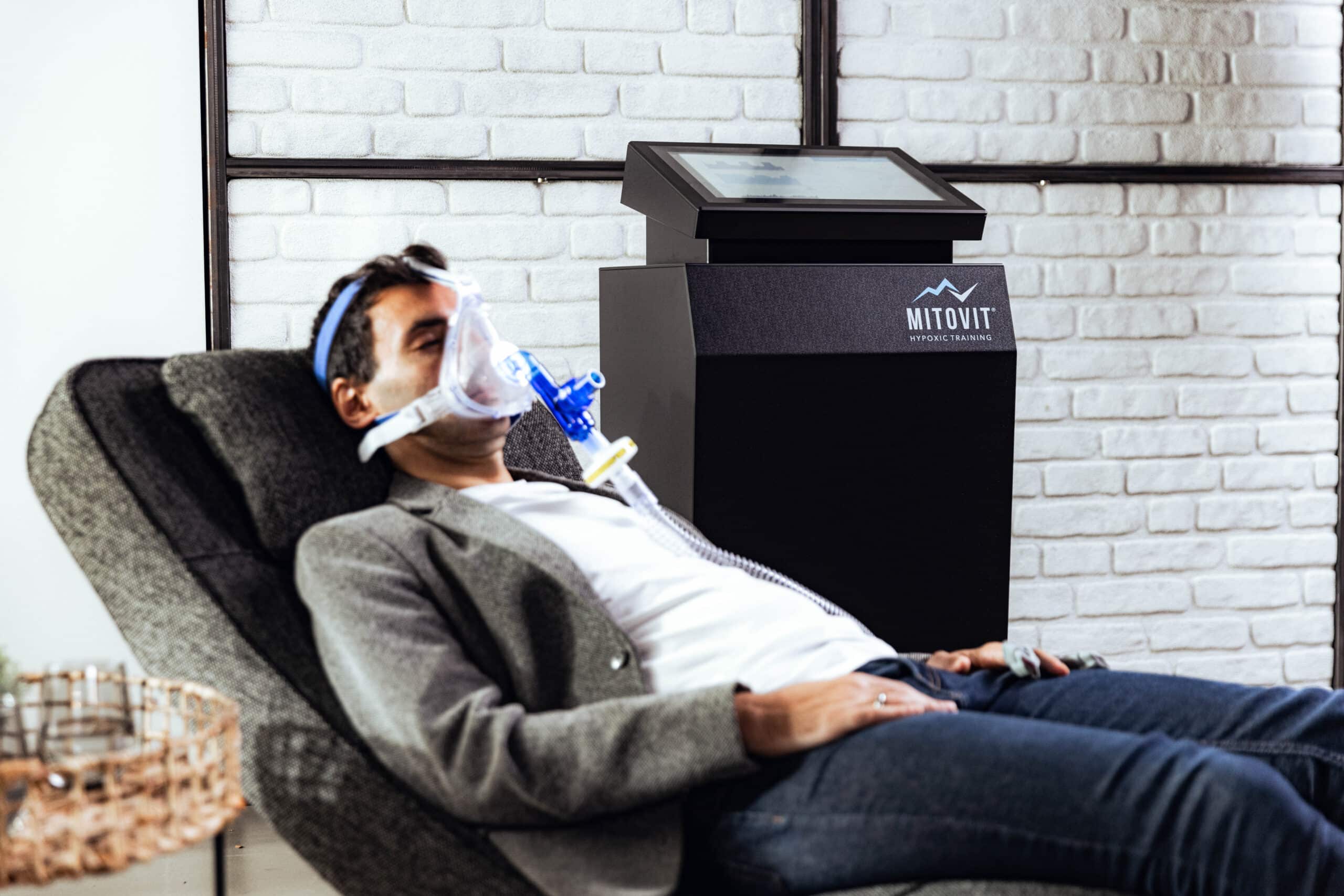 Eine Person liegt auf einem Stuhl und trägt eine durchsichtige Sauerstoffmaske, die mit einem Schlauch verbunden ist. In der Nähe steht ein "Mitovit Hypoxic Training"-Gerät mit einem Touchscreen, das für das IHHT Wolfenbüttel verwendet wird, vor einer weißen Backsteinwand.