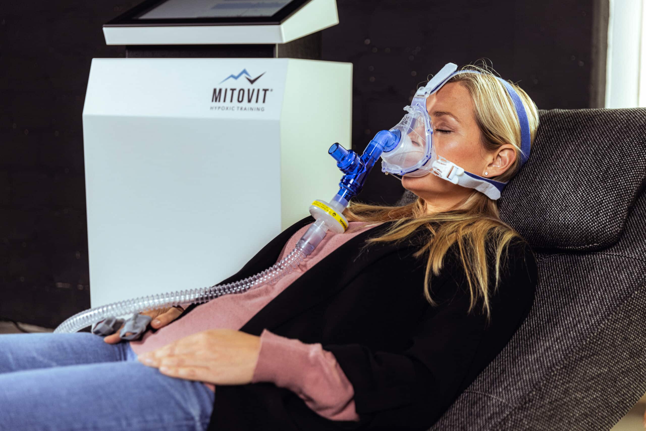 Eine Frau sitzt auf einem Liegestuhl und trägt eine Atemmaske, die an ein Gerät mit der Aufschrift "Mitovit Hypoxic Training" angeschlossen ist. Sie wirkt entspannt, hat die Augen geschlossen und erlebt das IHHT Wolfenbüttel, während ihre Hände ruhig auf ihrem Schoß ruhen.