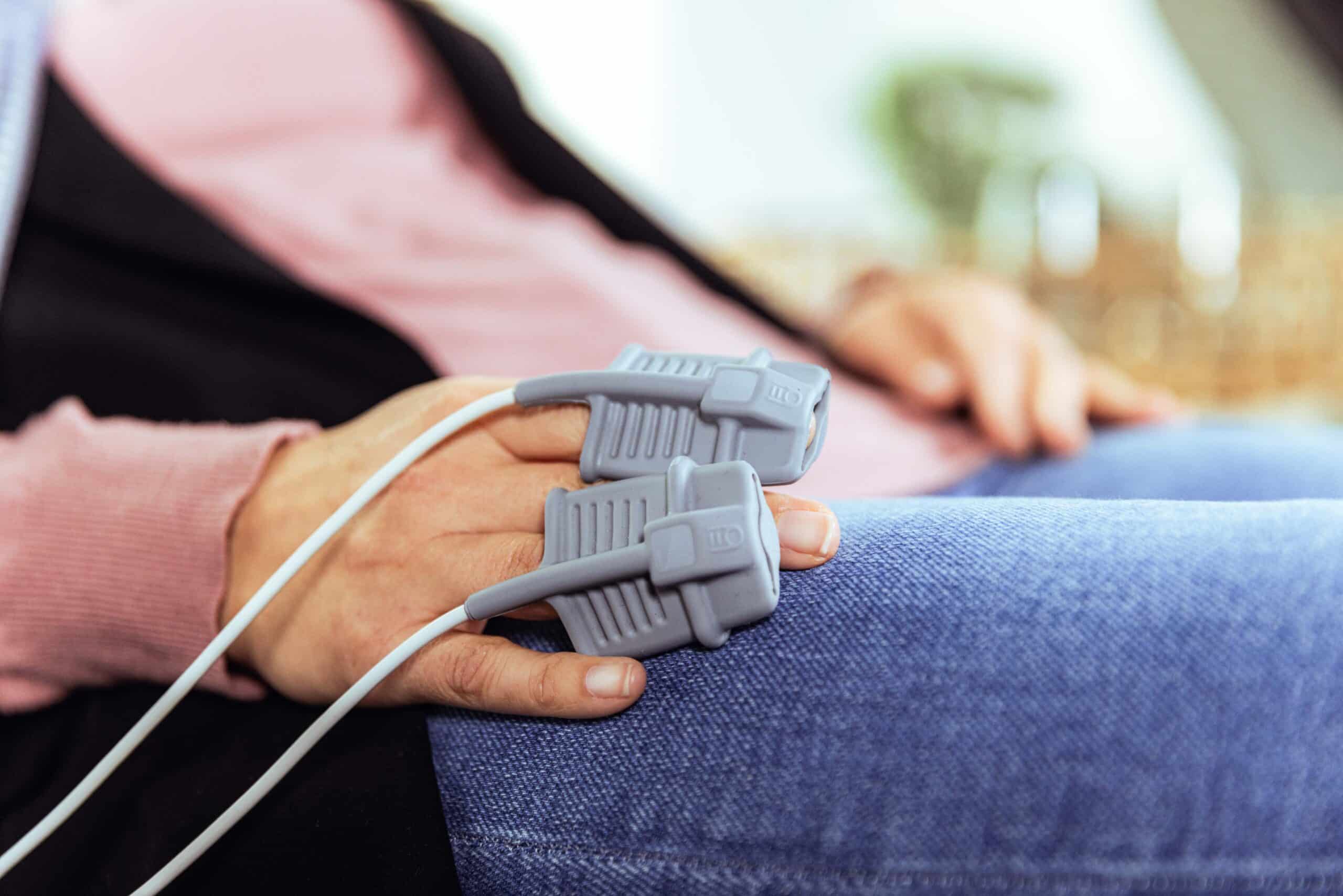 Während einer Sitzung des IHHT Wolfenbüttel ruht die Hand einer Person auf ihrem Schoß, an deren Fingern zwei medizinische Pulsoximeter-Sensoren mit weißen Drähten befestigt sind. Die Person trägt ein rosafarbenes langärmeliges Hemd und eine blaue Jeans.