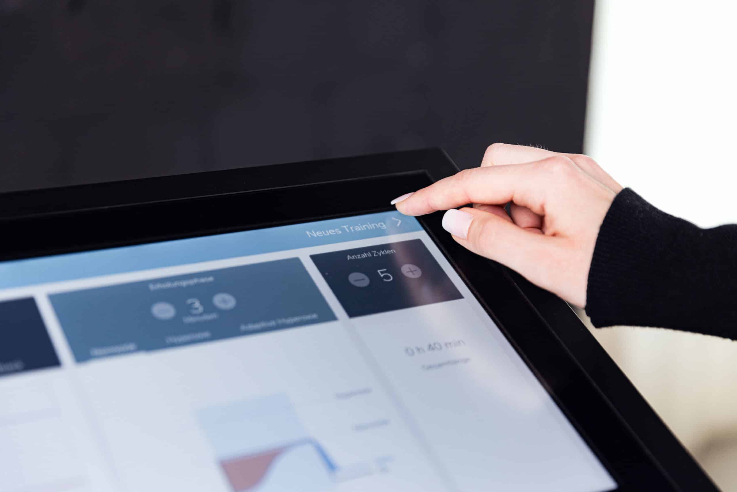 Die Hand einer Person berührt ein Touchscreen-Display, das Daten und Diagramme anzeigt, mit Optionen und der Aufschrift "Neues Training" oben rechts - Teil einer Sitzung des IHHT Wolfenbüttel.