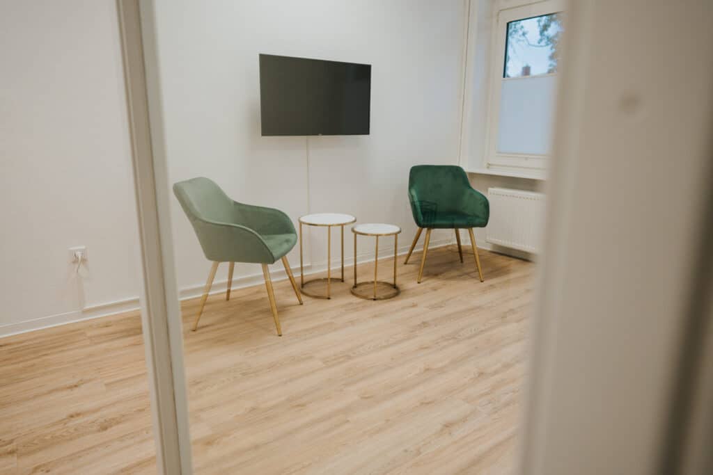 Ein kleiner Raum mit hellem Holzfußboden, zwei grünen Stühlen mit goldenen Beinen, zwei runden Beistelltischen und einem an der Wand befestigten Fernseher - perfekt für eine Neurologische Zweitmeinung in Wolfenbüttel. Das Foto ist von außerhalb des Raumes durch eine Türöffnung aufgenommen.