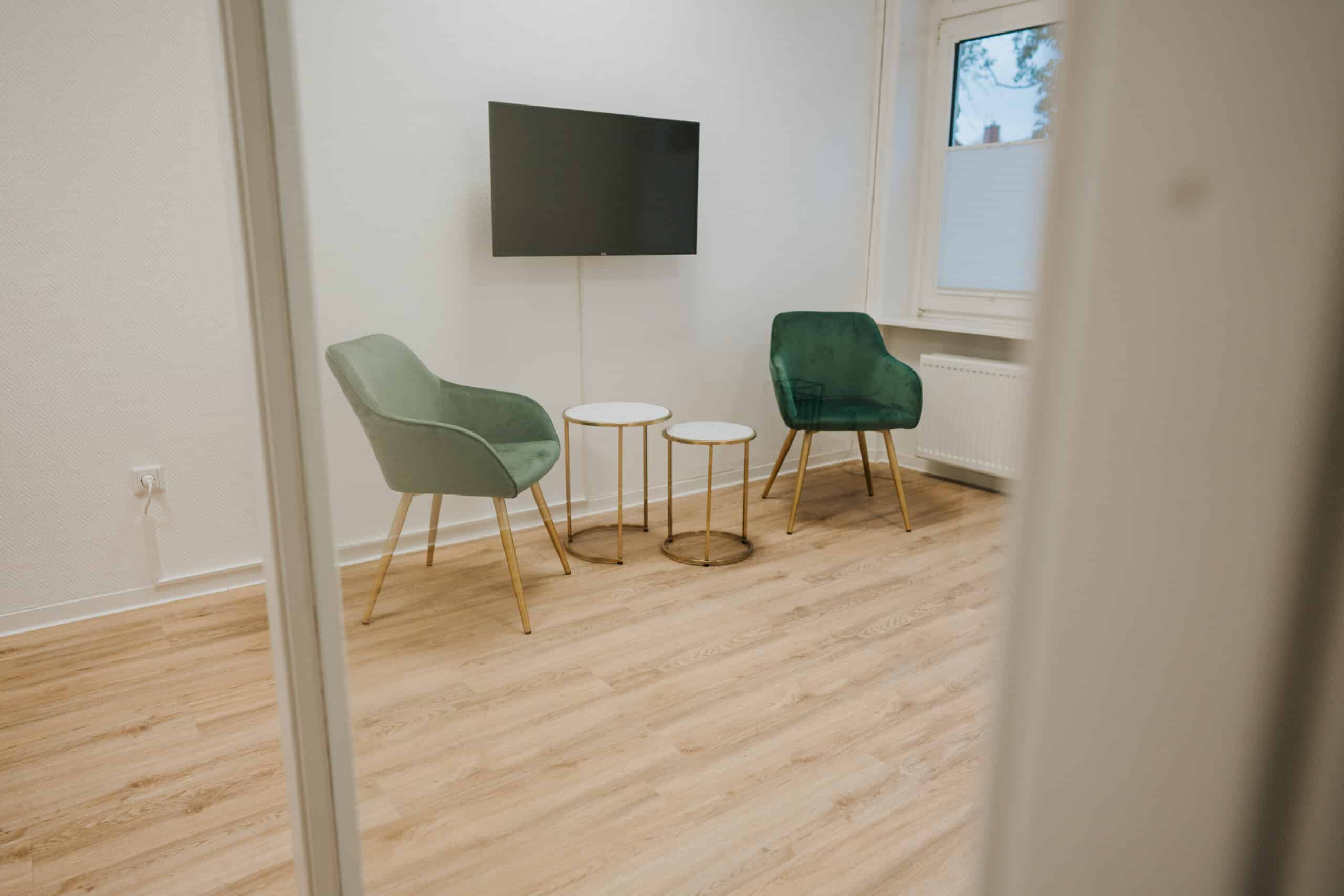 Ein kleiner Raum mit hellem Holzfußboden, zwei grünen Stühlen mit goldenen Beinen, zwei runden Beistelltischen und einem an der Wand befestigten Fernseher - perfekt für eine Neurologische Zweitmeinung in Wolfenbüttel. Das Foto ist von außerhalb des Raumes durch eine Türöffnung aufgenommen.