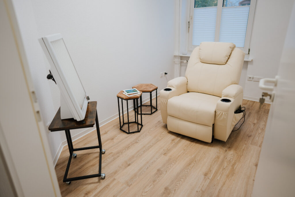 Ein kleiner Raum mit einem beigen Liegestuhl, zwei hölzernen Beistelltischen mit Büchern und einem Tablet sowie einem großen Spiegel auf einem Metallständer liegt auf einem hellen Holzboden in der Nähe eines Fensters - ideal für Sitzungen der Neurologischen Therapie Wolfenbüttel.