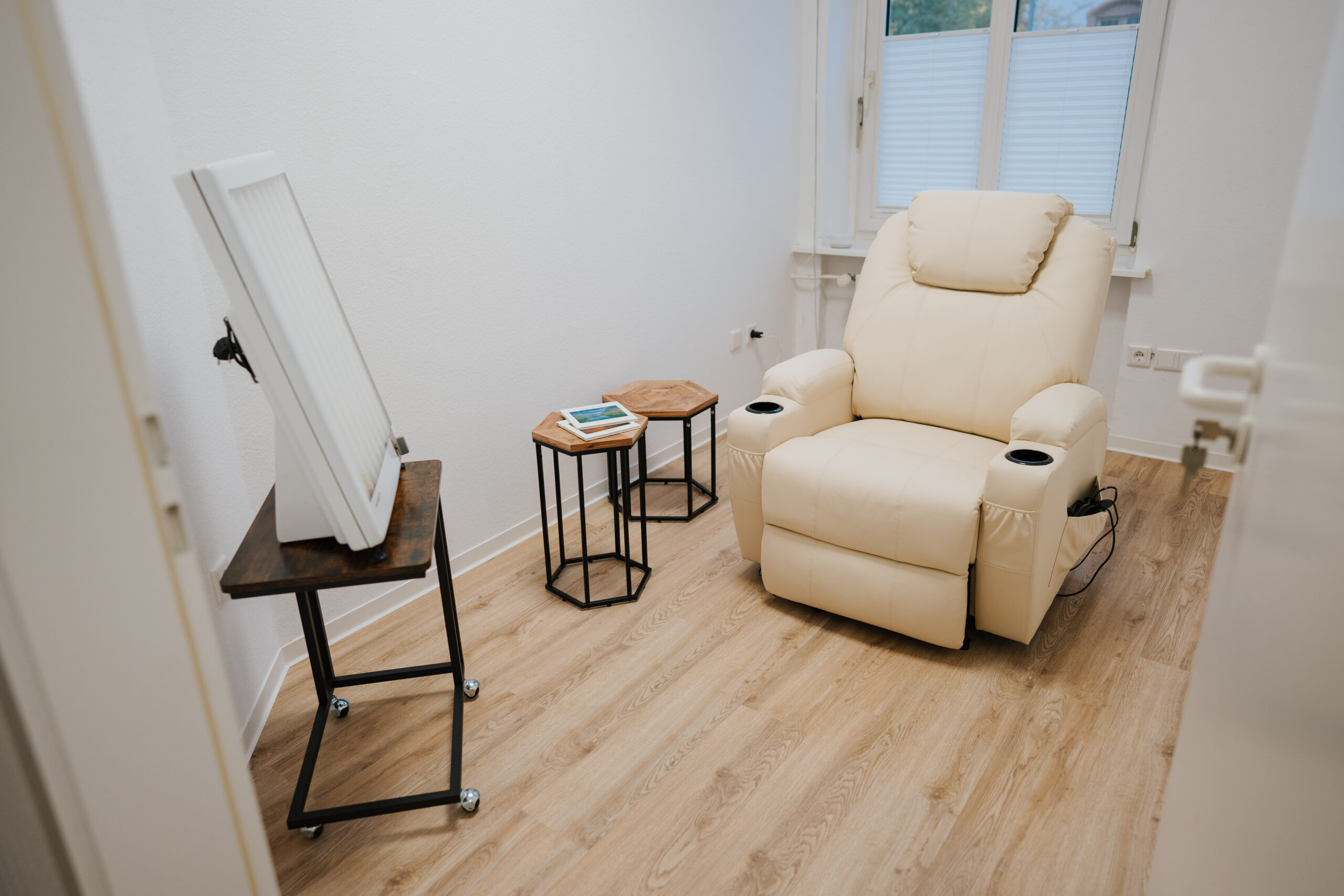 Ein kleiner Raum mit einem beigen Liegestuhl, zwei hölzernen Beistelltischen mit Büchern und einem Tablet sowie einem großen Spiegel auf einem Metallständer liegt auf einem hellen Holzboden in der Nähe eines Fensters - ideal für Sitzungen der Neurologischen Therapie Wolfenbüttel.