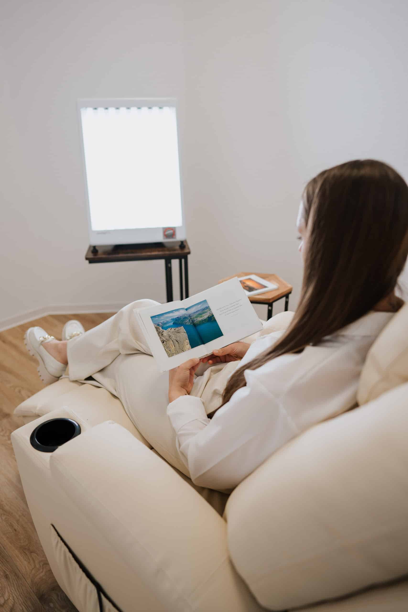 Eine Person mit langen braunen Haaren sitzt auf einer weißen Liege in der Neurologischen Praxis Wolfenbüttel und liest einen aufgeschlagenen Bildband. Auf einem kleinen Tisch daneben steht ein Tablet. Durch ein gut beleuchtetes Fenster im Hintergrund fällt natürliches Licht.