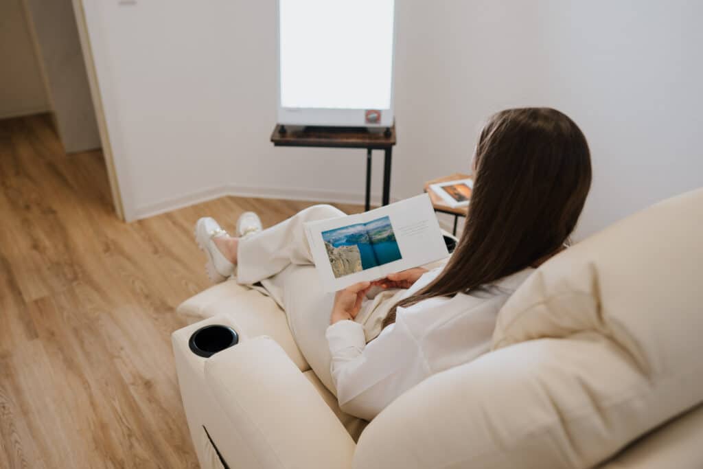 Eine Person mit langen braunen Haaren sitzt in einem cremefarbenen Liegestuhl und liest ein Buch oder eine Zeitschrift. In der Nähe steht ein Wolfenbütteler Neurofeedback-Gerät neben einer Lichttherapie-Lampe und einem kleinen Tisch mit Notebook in diesem aufgeräumten Raum mit Holzfußboden.