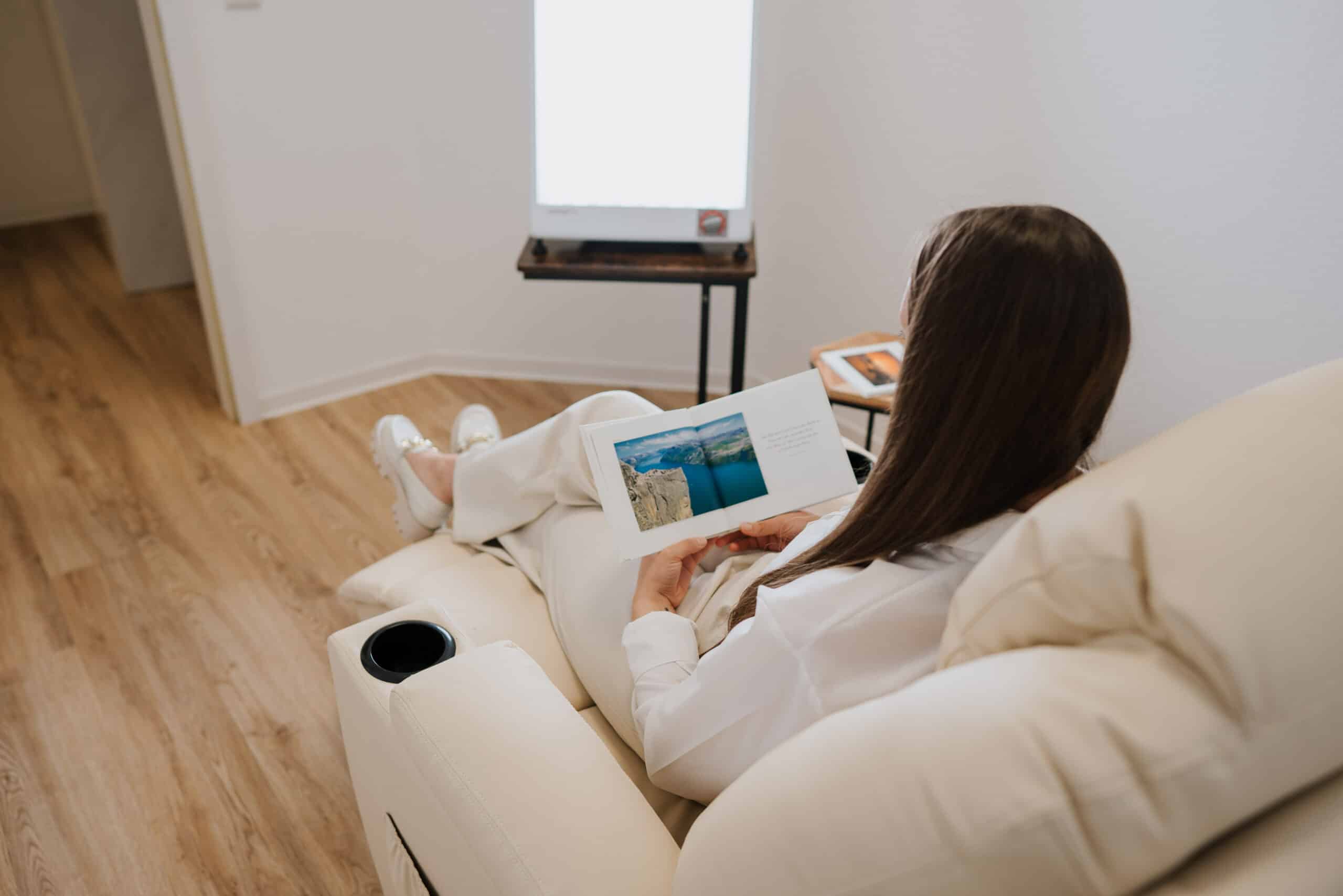 Eine Person mit langen braunen Haaren sitzt in einem cremefarbenen Liegestuhl und liest ein Buch oder eine Zeitschrift. In der Nähe steht ein Wolfenbütteler Neurofeedback-Gerät neben einer Lichttherapie-Lampe und einem kleinen Tisch mit Notebook in diesem aufgeräumten Raum mit Holzfußboden.