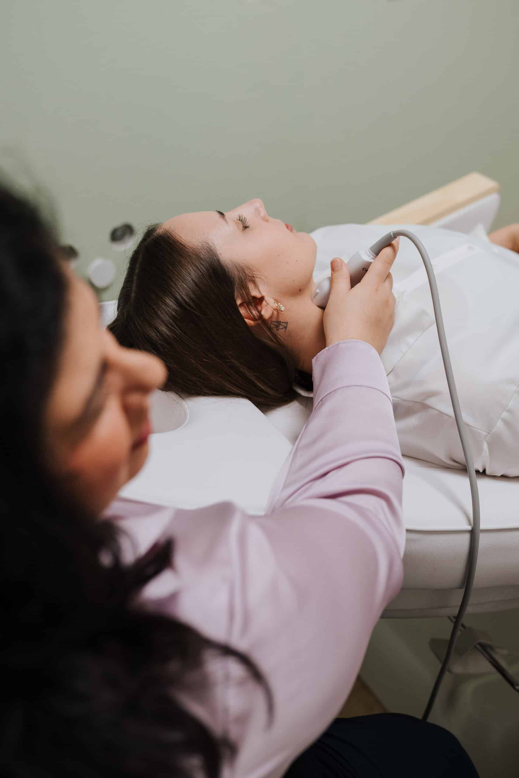 Eine Person liegt auf einem Untersuchungstisch, während eine andere Person ein medizinisches Handgerät am Hals trägt und möglicherweise eine Ultraschalluntersuchung oder einen Scan im Rahmen der Neurologischen Therapie Wolfenbüttel durchführt.