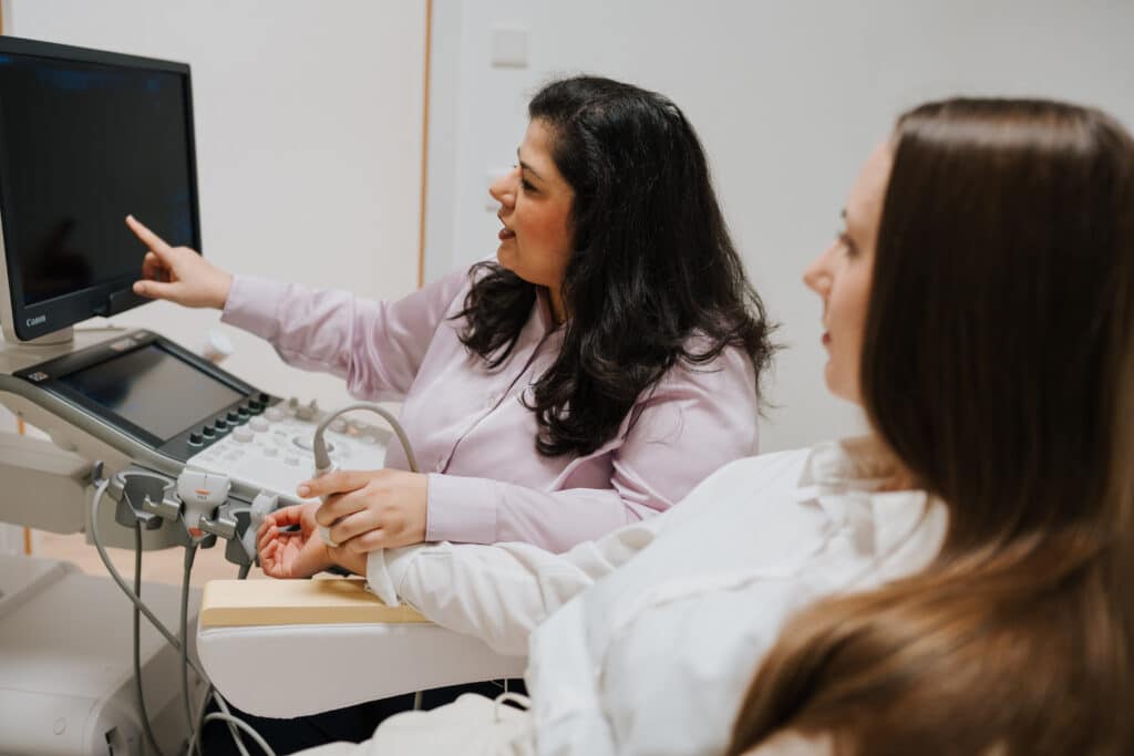 Eine medizinische Fachkraft zeigt auf einen Ultraschallmonitor, während eine andere Frau zusieht. Beide sitzen neben den medizinischen Geräten in einer klinischen Neurofeedback Wolfenbüttel Umgebung.
