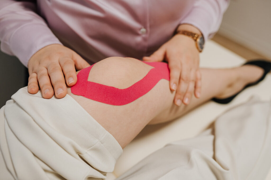 Eine Person, die eine helle Hose und ein rosafarbenes Hemd trägt, klebt sich ein rosafarbenes kinesiologisches Band um das Knie, während sie ihre Hände auf dem Bein ruhen lässt, möglicherweise nach einer Sitzung in der Neurologischen Therapie Wolfenbüttel.