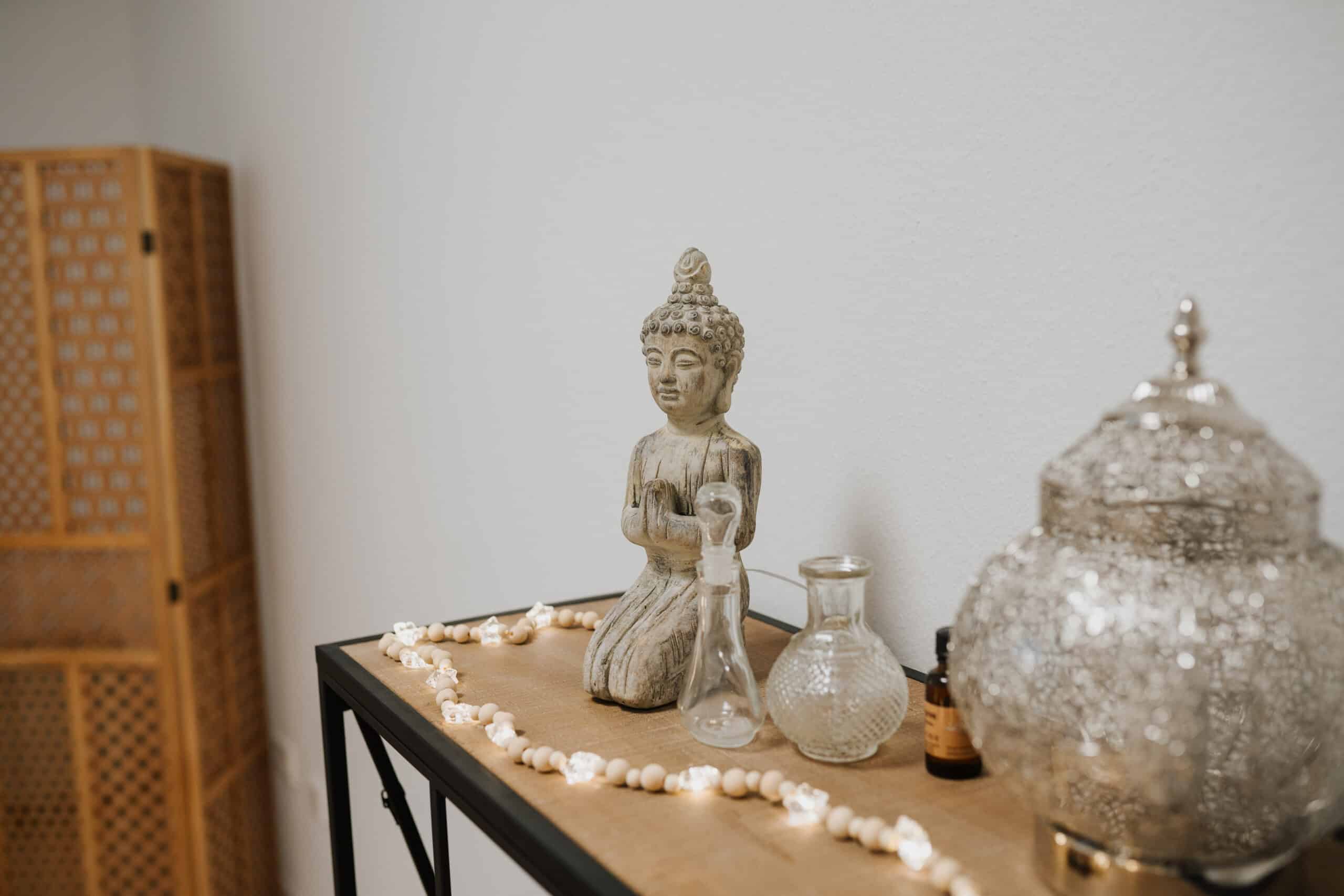 Auf einem Holztisch stehen eine Buddha-Statue, Glasvasen, ein dekoratives Gefäß, kleine Flaschen und ein Muschelstrang. Im Hintergrund vor einer schlichten weißen Wand steht ein Paravent, der an die Neurologische Zweitmeinung Wolfenbüttel erinnert.