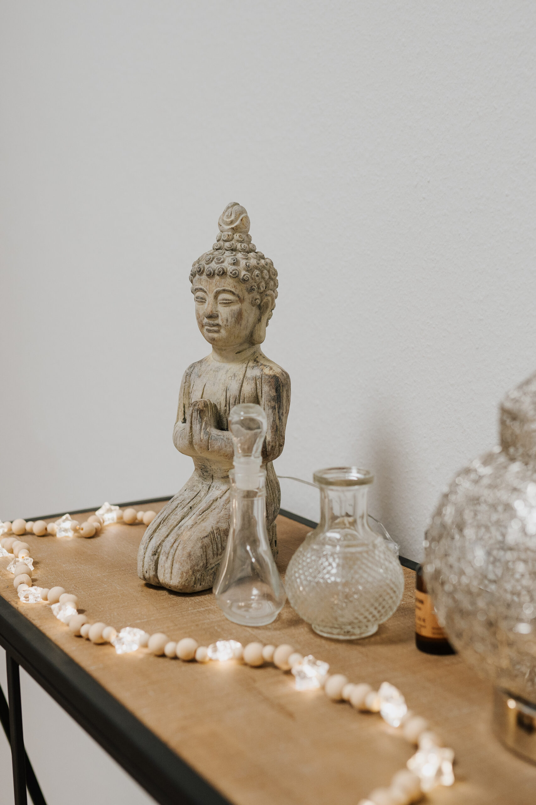 Eine kleine Buddha-Statue, Glasflaschen und eine Holzperlenkette mit Lichtern sind auf einer Holzfläche im beruhigenden Empfangsbereich der Neurologischen Praxis Wolfenbüttel vor einer schlichten weißen Wand angeordnet.