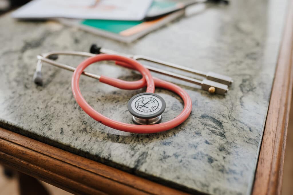 Ein rotes Stethoskop liegt auf einer marmornen Arbeitsplatte am Rand, im Hintergrund verschwimmen Papiere und ein grünes Notizbuch - ein einladender Rahmen für eine Sprechstunde der Neurologischen Zweitmeinung Wolfenbüttel.