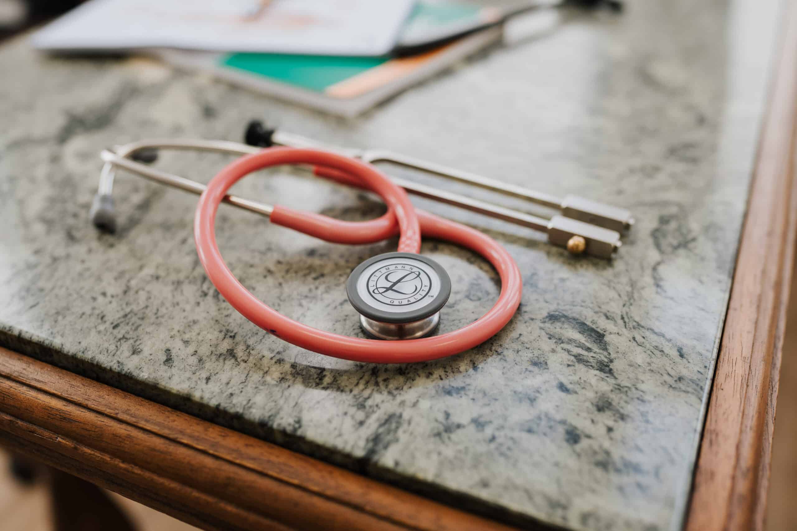 Ein rotes Stethoskop liegt auf einer marmornen Arbeitsplatte am Rand, im Hintergrund verschwimmen Papiere und ein grünes Notizbuch - ein einladender Rahmen für eine Sprechstunde der Neurologischen Zweitmeinung Wolfenbüttel.