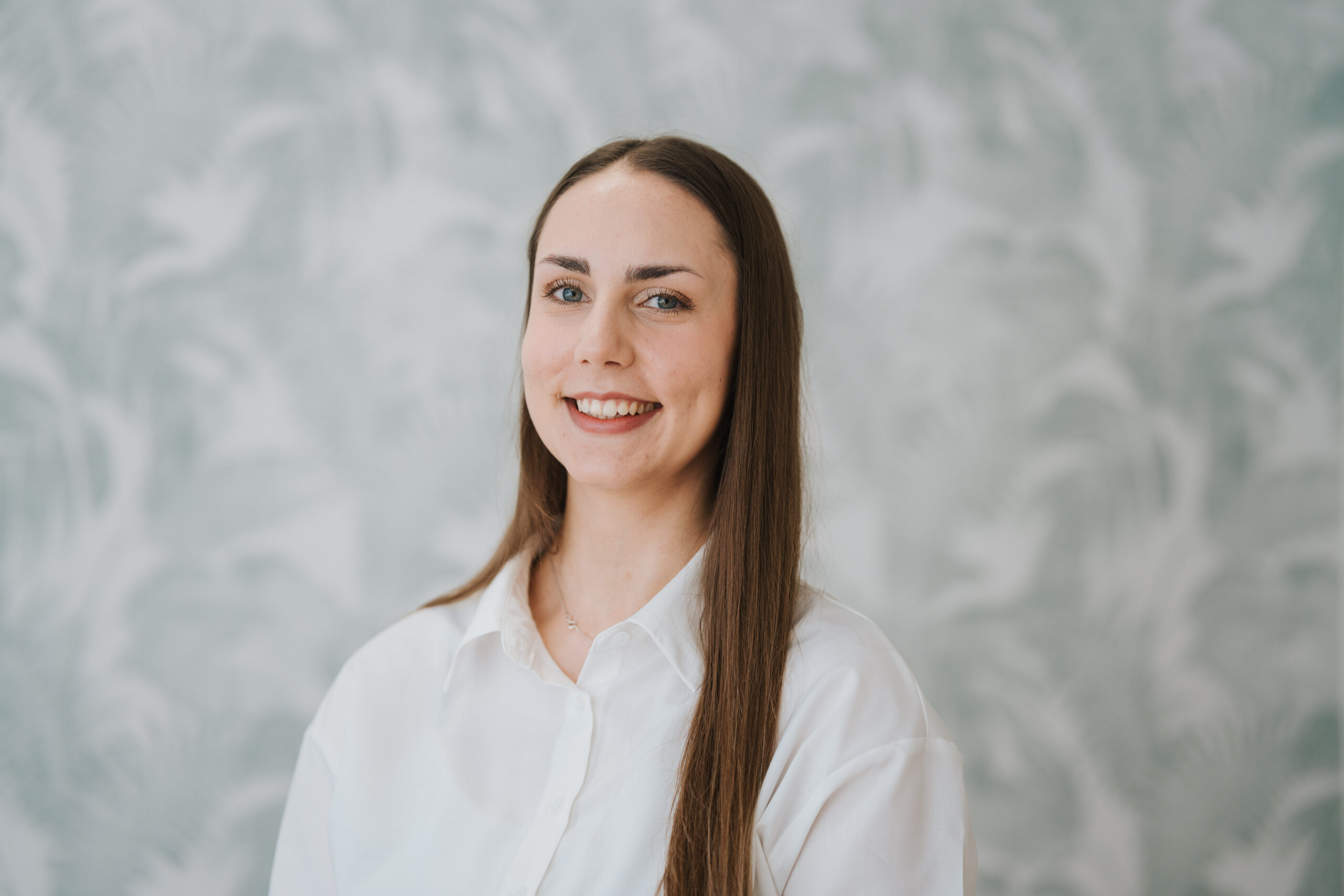 Eine Frau mit langen, glatten braunen Haaren und einem weißen Hemd steht lächelnd vor einem hellen, gemusterten Hintergrund in der Neurologischen Praxis Wolfenbüttel.