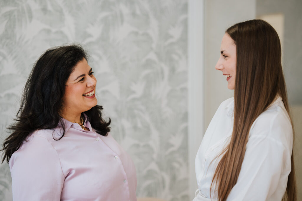 Zwei Frauen stehen sich lächelnd in einem Raum der Neurologischen Praxis Wolfenbüttel gegenüber und unterhalten sich vielleicht. Die eine hat langes dunkles Haar und trägt eine hellrosa Bluse, die andere hat glattes braunes Haar und ein weißes Hemd.