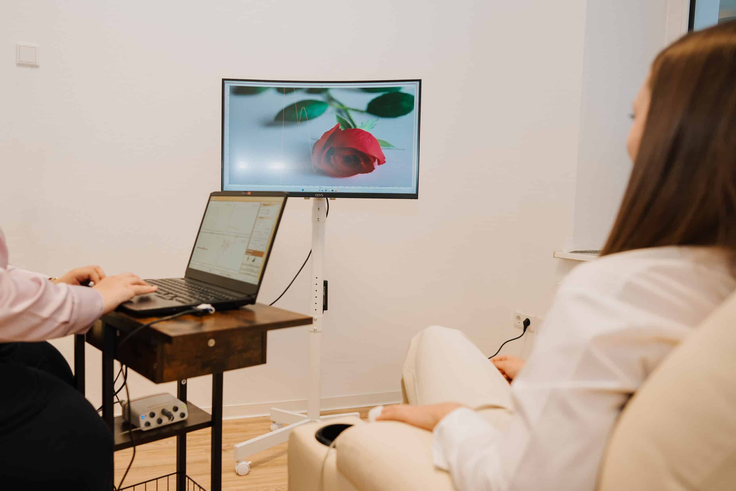 Zwei Personen sitzen in einem Büro, eine davon mit einem Laptop auf einem kleinen Schreibtisch. Auf einem Monitor ist die Nahaufnahme einer roten Rose zu sehen. Der helle Raum, der für Neurofeedback-Sitzungen in Wolfenbüttel vorgesehen ist, hat weiße Wände und eine minimalistische Einrichtung.
